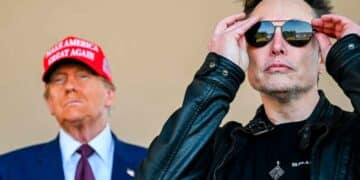 Primer plano de Elon Musk con chaqueta oscura y gafas de sol mientras, al fondo está Trump con una gorra con un mensaje propagandístico