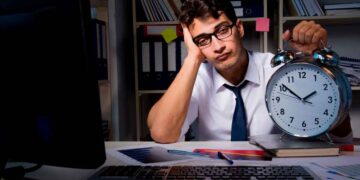 Trabajador de oficina cansado sostiene un gran reloj despertador, reflejando la necesidad de cambiar el horario laboral para conciliar
