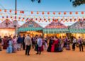 Casetas públicas de la Feria de Abril de Sevilla 2025 iluminadas al atardecer, con visitantes vestidos de flamenca disfrutando del ambiente