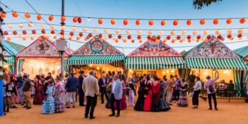 Casetas públicas de la Feria de Abril de Sevilla 2025 iluminadas al atardecer, con visitantes vestidos de flamenca disfrutando del ambiente