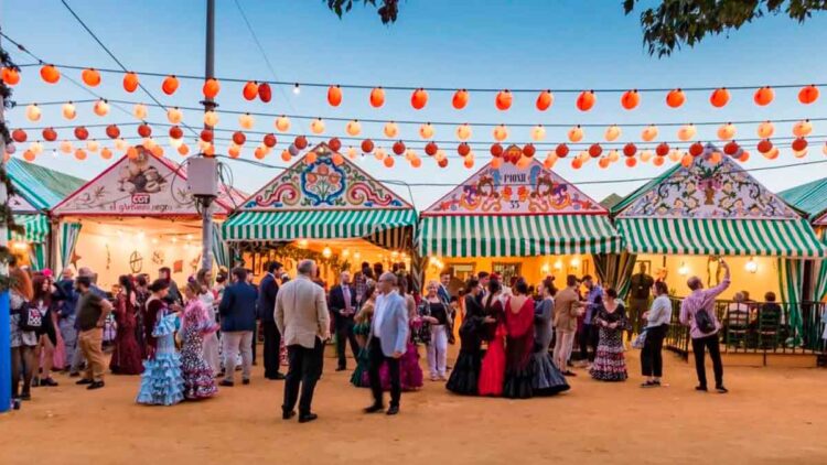 Casetas públicas de la Feria de Abril de Sevilla 2025 iluminadas al atardecer, con visitantes vestidos de flamenca disfrutando del ambiente