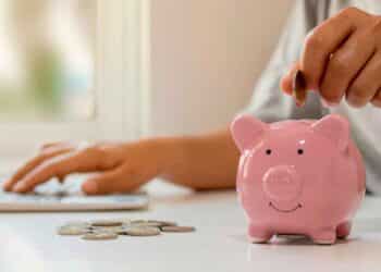 Persona introduciendo una moneda en una hucha rosa mientras usa un portátil, simbolizando el ahorro en depósitos bancarios a corto plazo