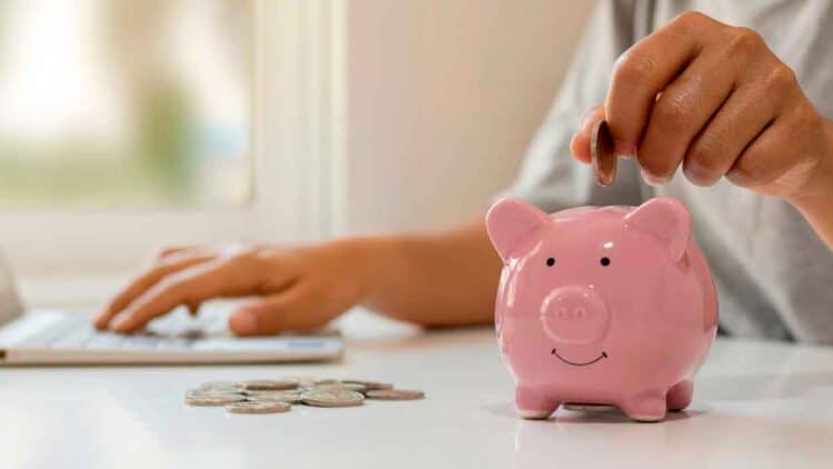 Persona introduciendo una moneda en una hucha rosa mientras usa un portátil, simbolizando el ahorro en depósitos bancarios a corto plazo