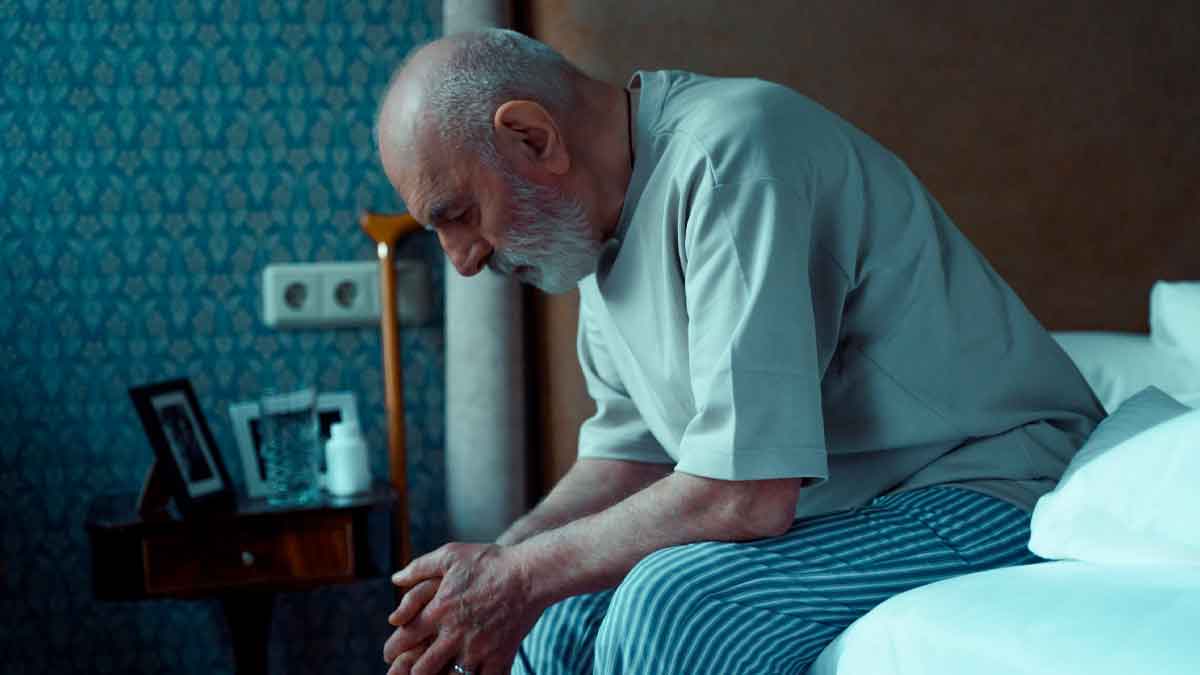 Hombre mayor cabizbajo sentado en la cama, vestido con pijama, en una habitación con decoración clásica