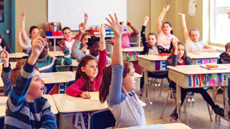 Niños y niñas levantando la mano en clase, ejemplo de alumnado que puede beneficiarse de las becas 2025/26 de apoyo educativo