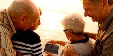 Grupo de jubilados consultando una tablet al atardecer junto al mar sobre la pensión máxima en Europa, EE. UU. y Canadá