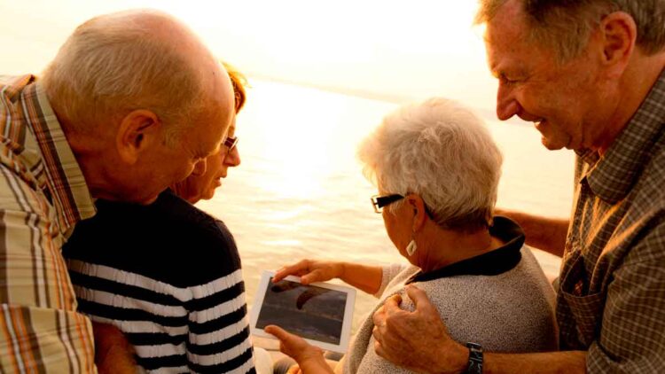 Grupo de jubilados consultando una tablet al atardecer junto al mar sobre la pensión máxima en Europa, EE. UU. y Canadá