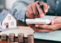Persona usando una calculadora junto a monedas y una maqueta de casa para calcular la devolución de gastos hipotecarios antes de que prescriba el plazo