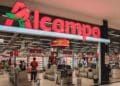 Entrada de un supermercado Alcampo con su logo iluminado; la cadena aplica un ERE con 565 despidos e indemnización de 35 días por año