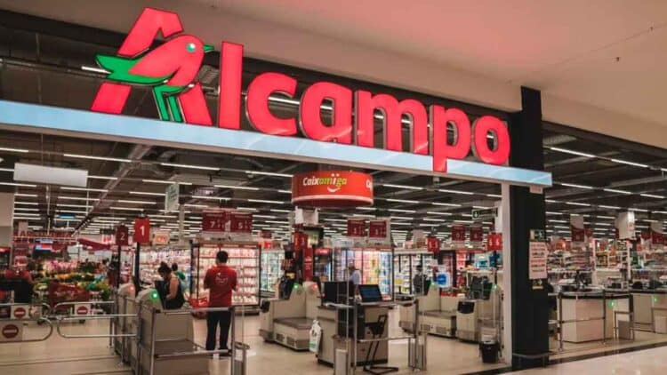 Entrada de un supermercado Alcampo con su logo iluminado; la cadena aplica un ERE con 565 despidos e indemnización de 35 días por año