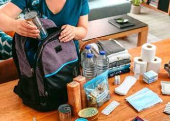 Mujer colocando latas y agua en una mochila de kit de supervivencia sobre la mesa, rodeada de suministros básicos ante un posible apagón en España