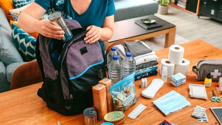 Mujer colocando latas y agua en una mochila de kit de supervivencia sobre la mesa, rodeada de suministros básicos ante un posible apagón en España