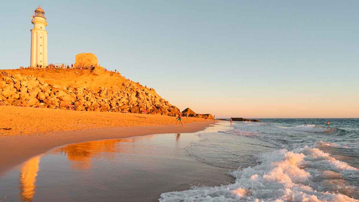 Playa del Faro de Trafalgar al atardecer con arena dorada, faro y olas; zona peligrosa sin socorristas y con baño prohibido