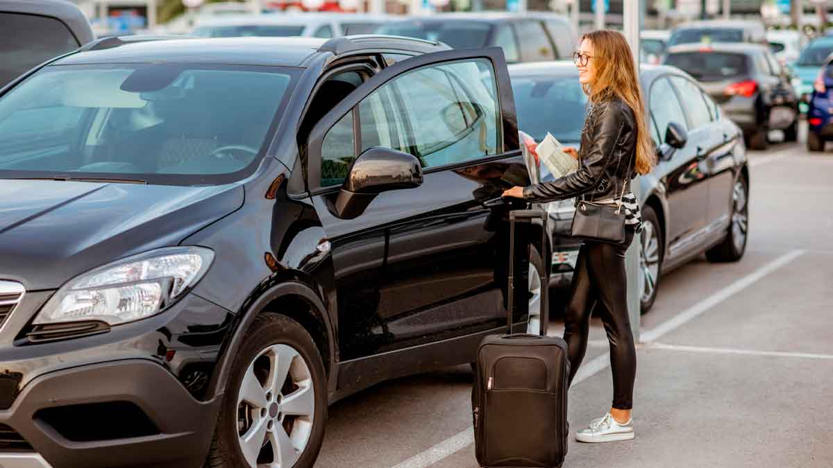 Mujer con maleta toma coche de alquiler negro en parking siguiendo consejos de Consumo para evitar abusos