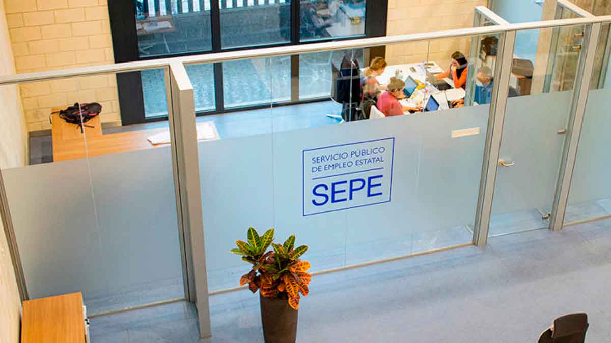 Interior de una oficina del SEPE donde empleados tramitan prestaciones por desempleo, con el logo visible en la mampara de cristal