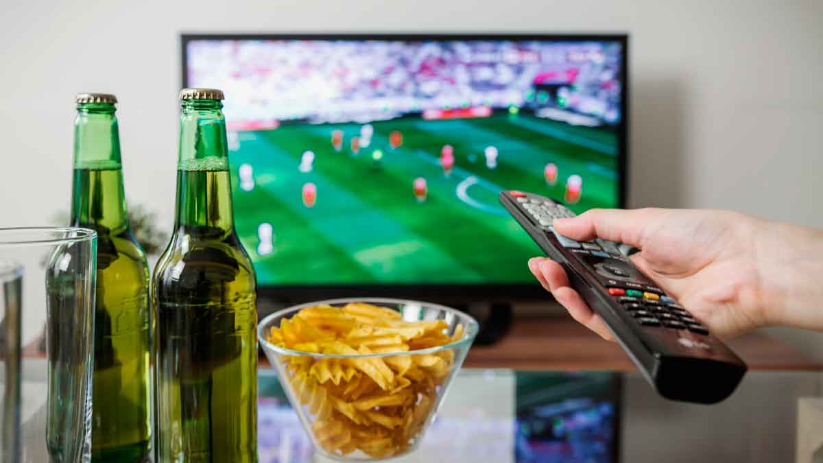 Partido de fútbol temporada 2025-2026 en televisión, mano con mando a distancia, cervezas y aperitivo sobre la mesa en un salón