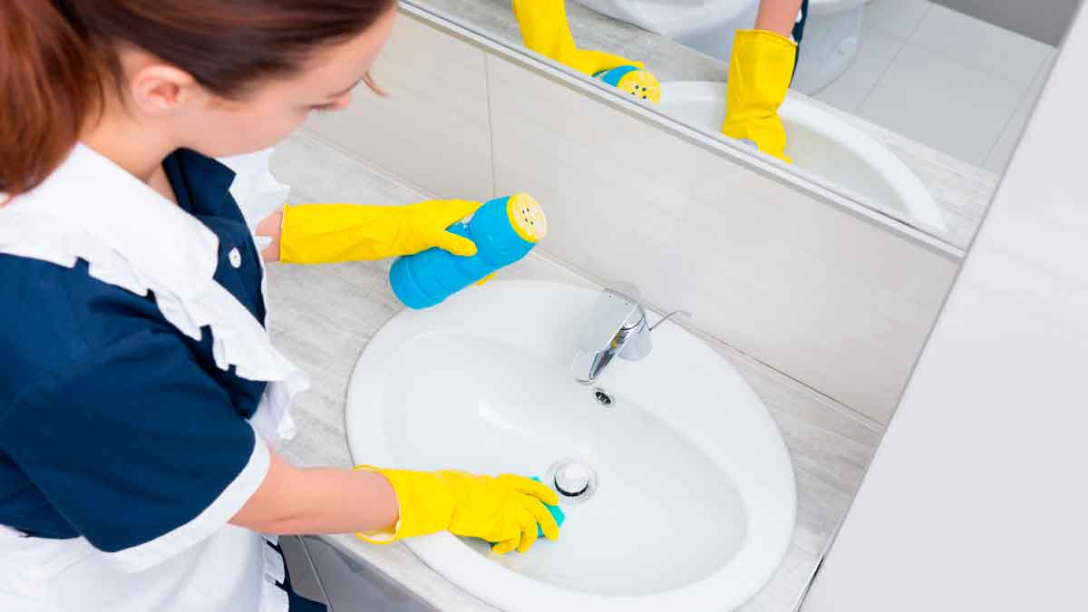 Empleada del hogar limpiando un lavabo con guantes y productos de limpieza