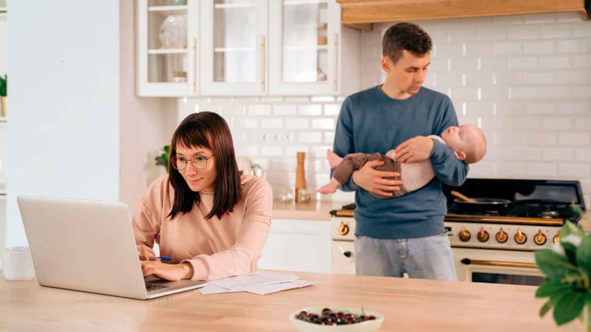 Trabajadora usando portátil en casa mientras su pareja cuida al bebé, ejemplo de permisos retribuidos por conciliación laboral