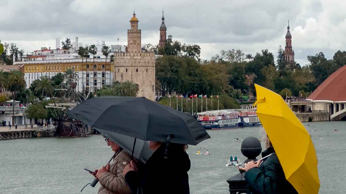 Personas con paraguas bajo la lluvia en Sevilla junto a la Torre del Oro durante un episodio de inestabilidad meteorológica