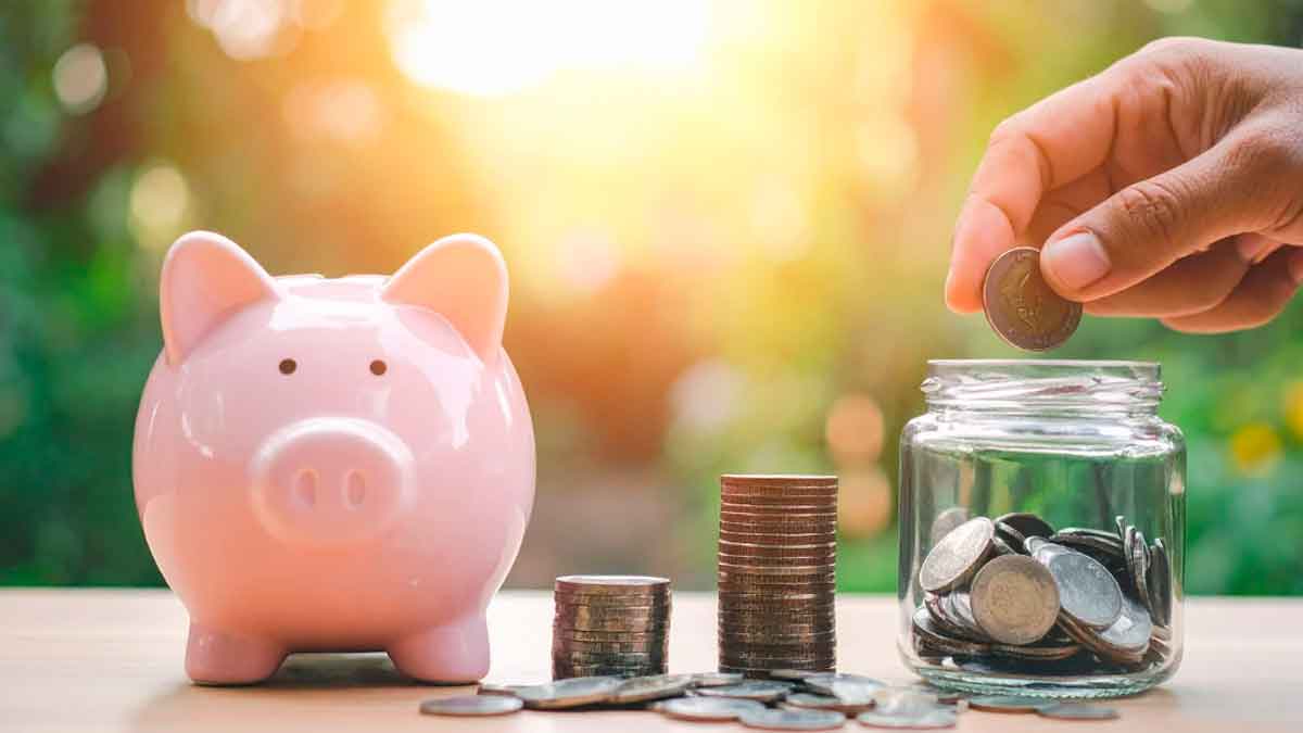 Hucha rosa y monedas apiladas junto a un tarro de ahorro con una mano introduciendo una moneda