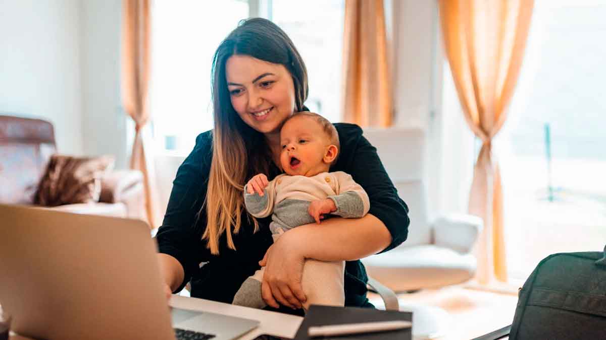 Madre trabajando con el ordenador mientras sostiene a su bebé en brazos en casa