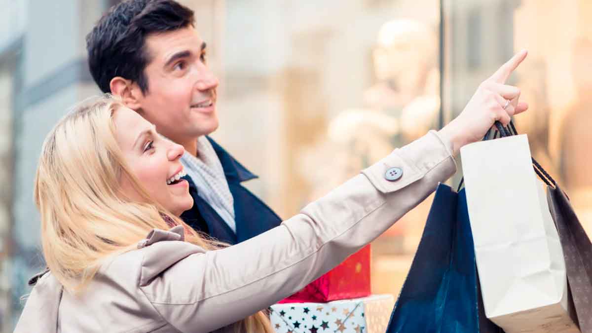 Pareja joven realizando compras navideñas mientras observa escaparates con bolsas y regalos
