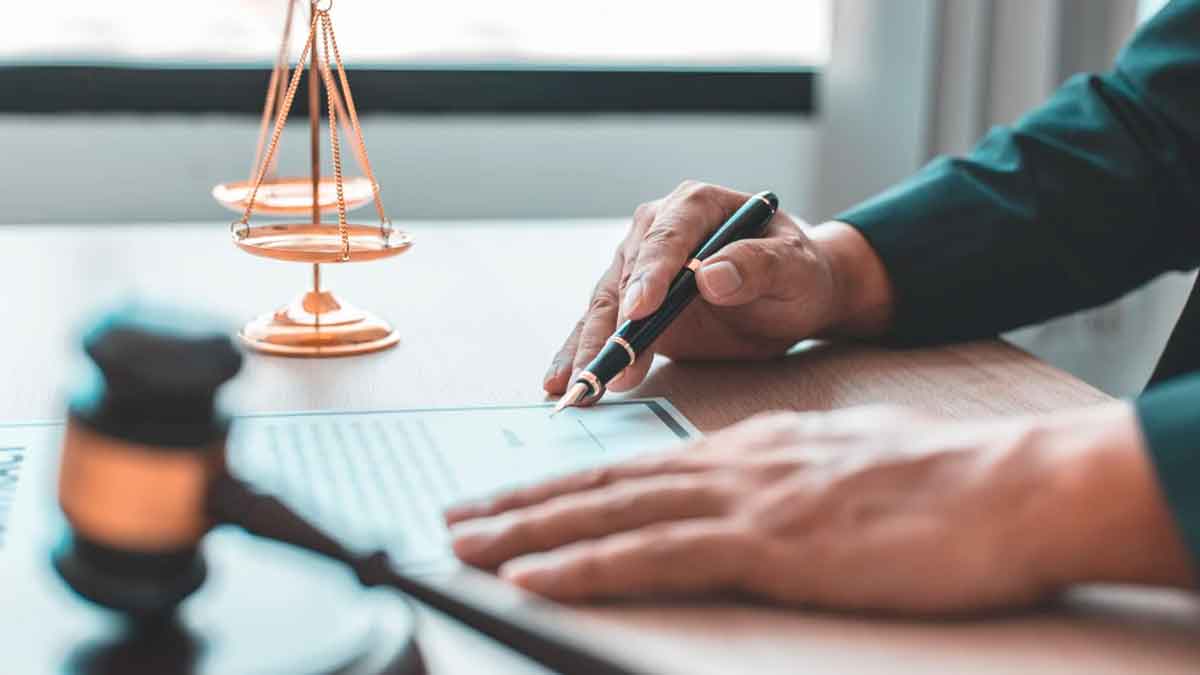 Manos firmando un documento jurídico junto a una balanza y un mazo de juez en una mesa